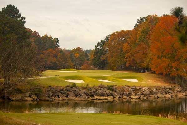 A scenic golf course with lush green fairways, sand traps, surrounded by autumn-colored trees and a calm water body.