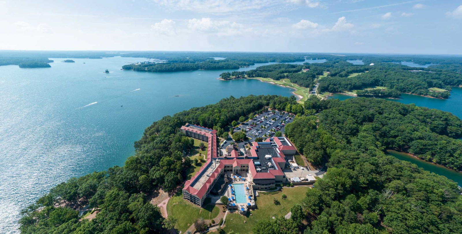 An aerial view of a large resort complex surrounded by lush greenery on the edge of a lake with several boats visible on the water.