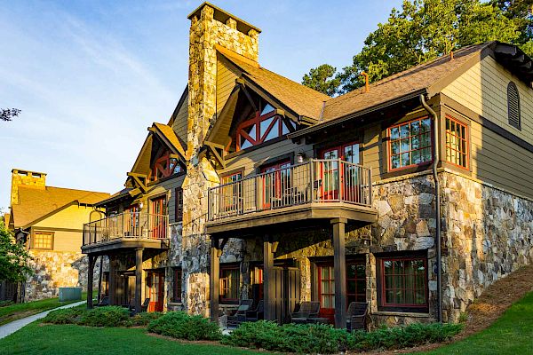 A large, rustic stone and wood house with two stories, balconies, and surrounded by greenery in a serene setting, likely in a rural area, is shown.