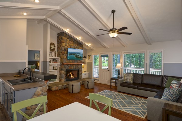 A cozy living room with a stone fireplace, large windows, a brown leather sofa, and a ceiling fan, blending comfort and nature.
