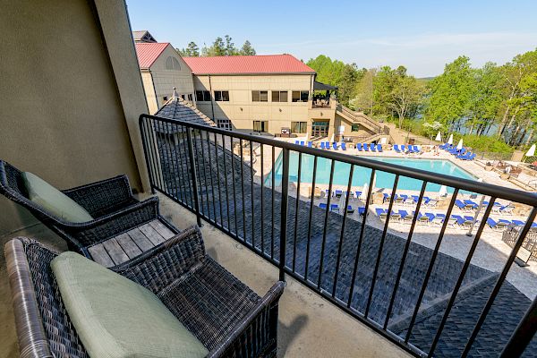 View from a balcony with two wicker chairs and a wrought iron railing, overlooking a resort pool area with sun loungers and a two-story building.