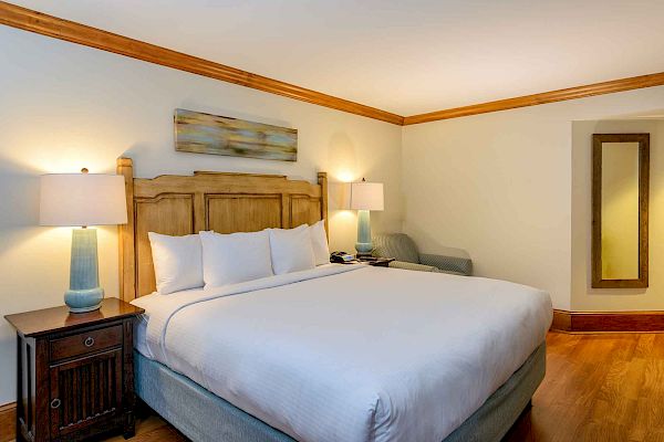 A well-lit bedroom featuring a king-sized bed with white bedding, bedside tables with lamps, a wall mirror, and a piece of abstract art.