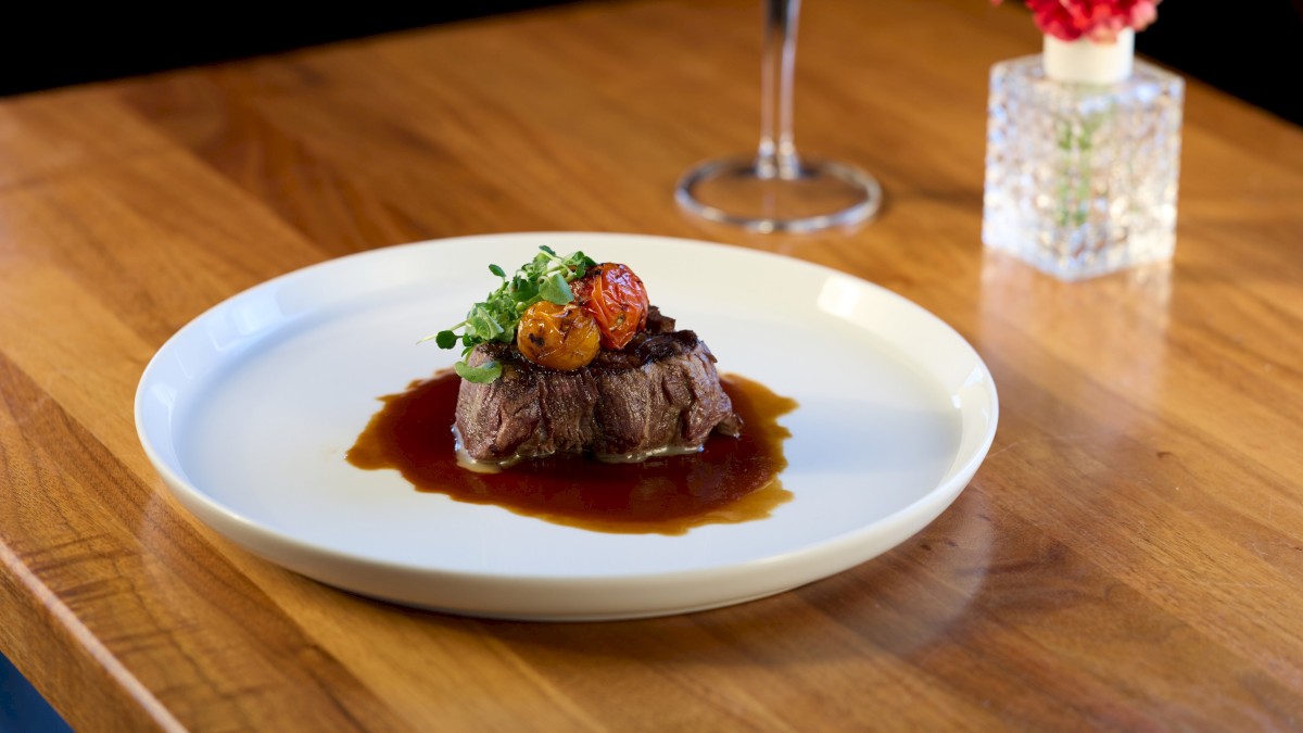 A plated upscale steak with sauce, garnished with greens and a small roasted vegetable, on a white plate at a wooden table.