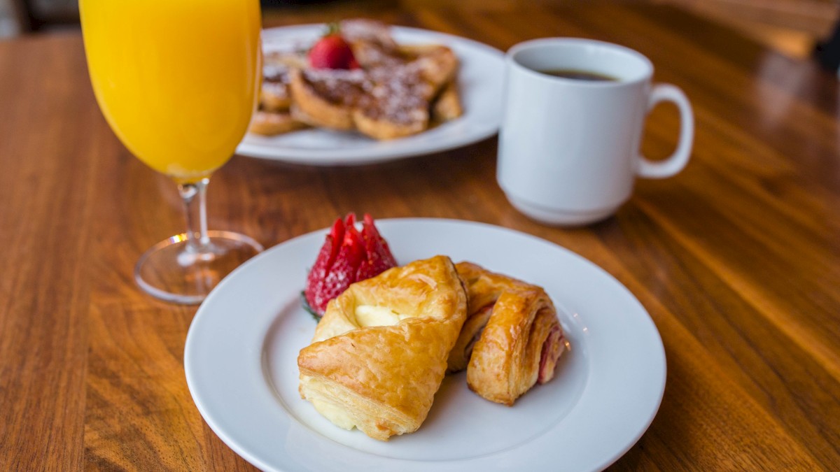 A breakfast plate with flaky pastries and a strawberry on a white plate, plus a glass of orange juice, a coffee mug, and a blurred pastry plate in the background.