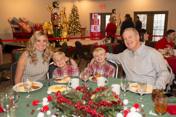 A family of four sits at a festive holiday table with greenery and berries, smiling for the camera in a decorated banquet hall.