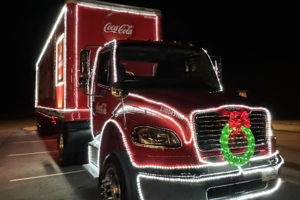 A Coca-Cola truck decorated with festive lights and a wreath is parked at night, shining brightly against the darkness.