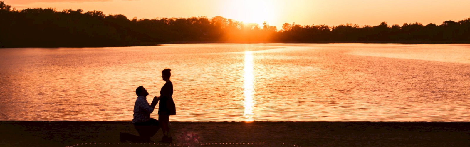 A couple is silhouetted against a sunset by a lake, with one person kneeling, surrounded by candlelight on the sand.