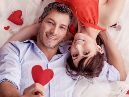 A couple lying down, smiling at the camera, holding a red heart shape. They are surrounded by white fabric and additional red heart shapes.