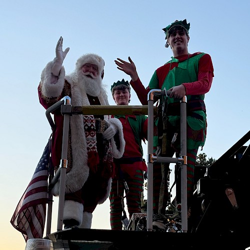 Santa and two people dressed as elves are standing on a decorated fire truck, waving.