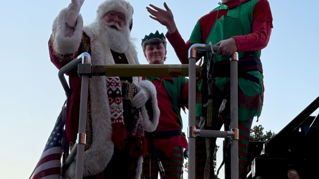 Santa and two people dressed as elves are standing on a decorated fire truck, waving.