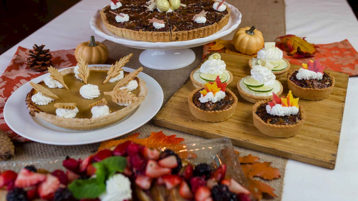 A festive autumn dessert spread with pies, tarts, and berry toppings, decorated with fall leaves, pumpkins, and pinecones.
