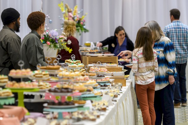 People browse a long dessert table at a buffet, selecting treats from an array of colorful cakes, cupcakes, and pastries.