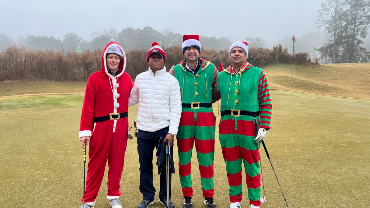 Four people dressed as Santa and elves stand on a golf course, posing together on a cloudy day.