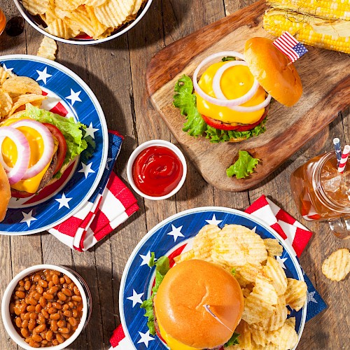 American-style burgers, potato chips, watermelon, baked beans, and soft drinks are arranged on a rustic wooden table for a casual meal.