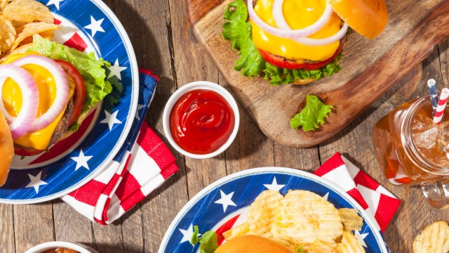 American-style burgers, potato chips, watermelon, baked beans, and soft drinks are arranged on a rustic wooden table for a casual meal.