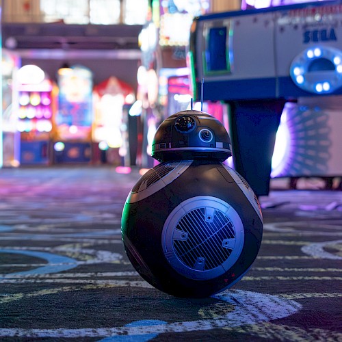 A BB-8 style robot is on a patterned floor in an arcade filled with colorful lights and games.