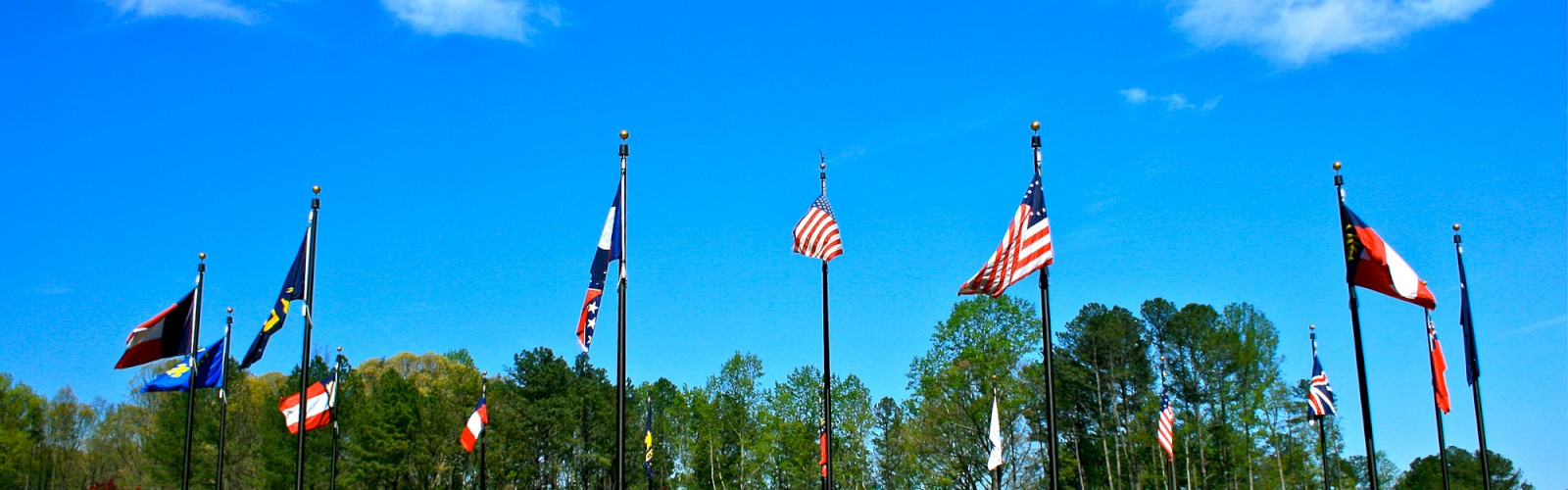 A roundabout with multiple flagpoles displaying various national and state flags set against a bright blue sky with scattered clouds.