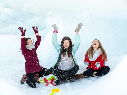 Three people are sitting in the snow, joyfully throwing snow into the air. They are dressed in winter clothing, including scarves and gloves.