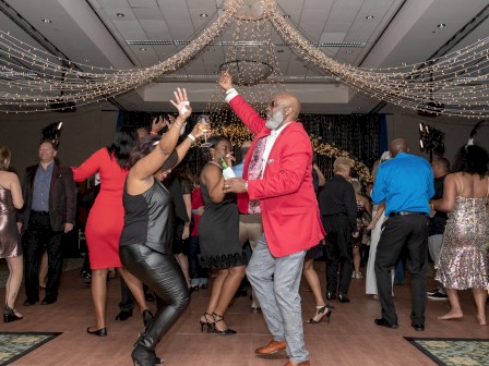 People are dancing and celebrating at an indoor party under decorative hanging lights, with a variety of outfits and lively expressions.