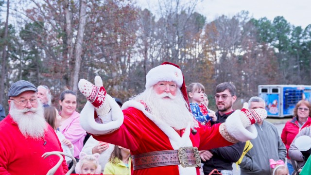 A cheerful Santa waves to a crowd in a park, with kids and adults gathered, festive outfits and a cozy holiday vibe. Ending this sentence.