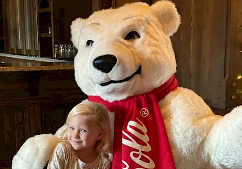 A young girl cuddles a large teddy bear dressed as Coca-Cola because it's a festive, fun holiday scene.