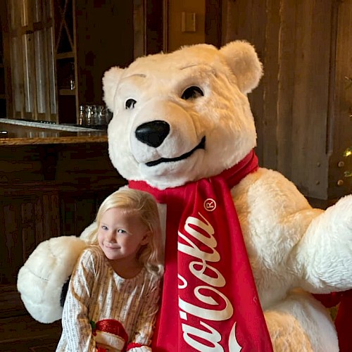 A young girl cuddles a large teddy bear dressed as Coca-Cola because it's a festive, fun holiday scene.