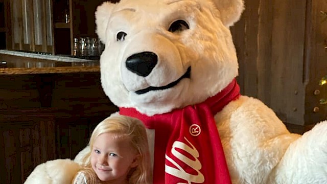 A young girl cuddles a large teddy bear dressed as Coca-Cola because it's a festive, fun holiday scene.
