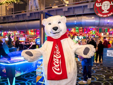 A person in a polar bear costume with a Coca-Cola scarf at a festive, colorful venue with holiday decorations and people enjoying the event.