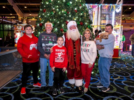 A family gathers with Santa Claus under a decorated Christmas tree at an indoor amusement park, celebrating the holiday season.