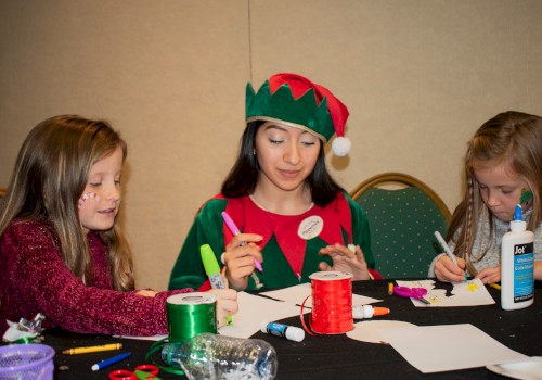 A woman dressed as an elf with two young girls are crafting with glue, scissors, markers, and ribbons at a table.