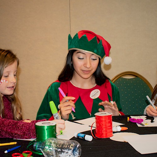 A woman dressed as an elf with two young girls are crafting with glue, scissors, markers, and ribbons at a table.