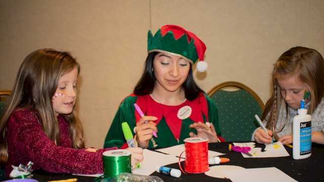 A woman dressed as an elf with two young girls are crafting with glue, scissors, markers, and ribbons at a table.