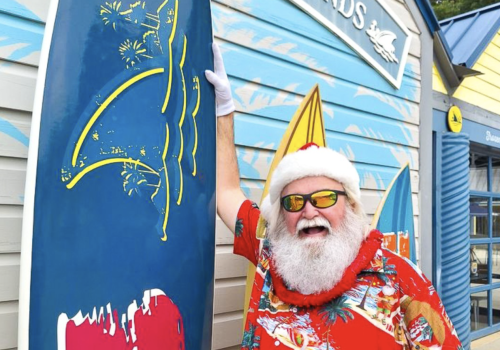 A cheerful man dressed in a festive Hawaiian shirt and Santa hat, with sunglasses, celebrating at a surf shop called 