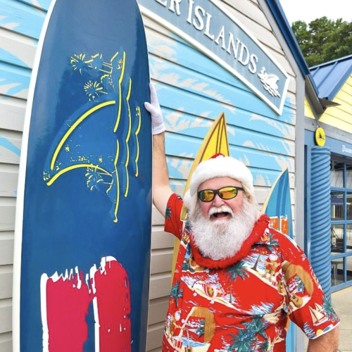 A cheerful man dressed in a festive Hawaiian shirt and Santa hat, with sunglasses, celebrating at a surf shop called 