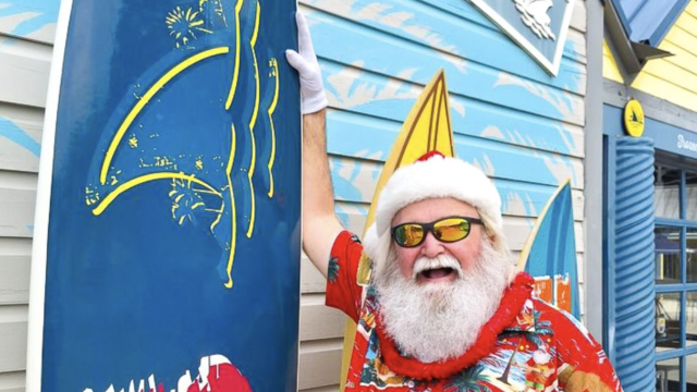 A cheerful man dressed in a festive Hawaiian shirt and Santa hat, with sunglasses, celebrating at a surf shop called 