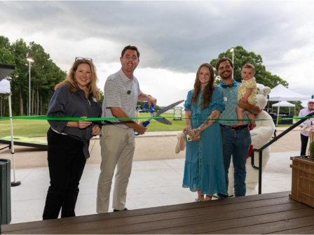 A group of five people, including a child, participate in a ribbon-cutting ceremony outdoors on a cloudy day.