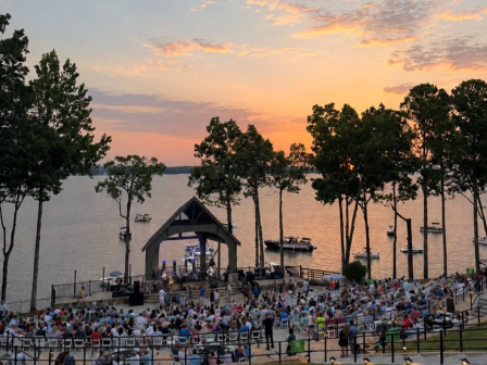 An outdoor concert takes place by the water at sunset, with a large audience seated, and boats floating on the lake nearby.