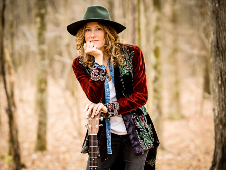 A woman wearing a hat and colorful jacket, holding a guitar, stands in a forest path.