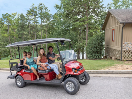 A family of four rides in a red golf cart on a quiet, tree-lined residential street near a house with a wooden exterior.