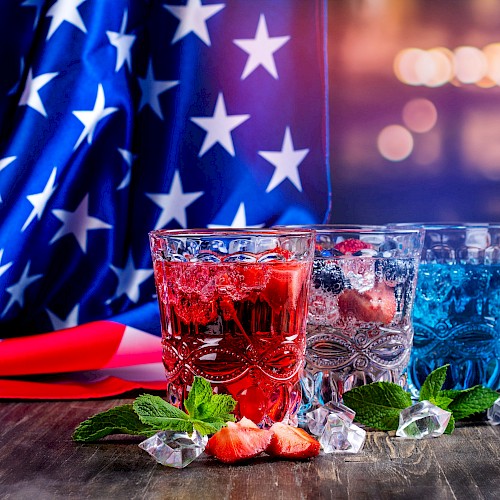Colorful patriotic drinks with ice, strawberries, and mint leaves are in front of an American flag for a celebration.