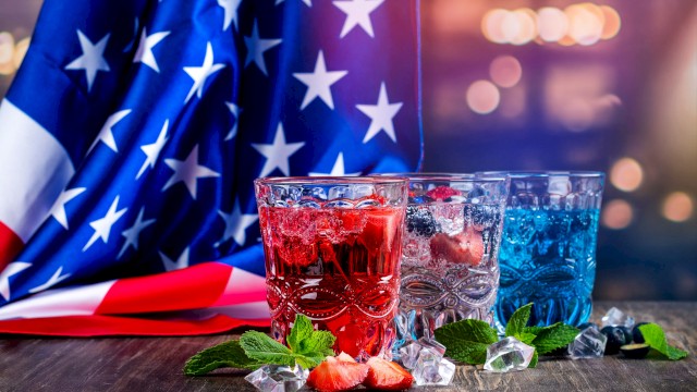 Colorful patriotic drinks with ice, strawberries, and mint leaves are in front of an American flag for a celebration.