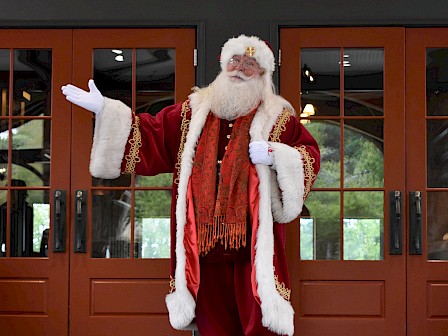 A person in a Santa Claus outfit stands on a stone floor in front of doors, gesturing with one arm raised.