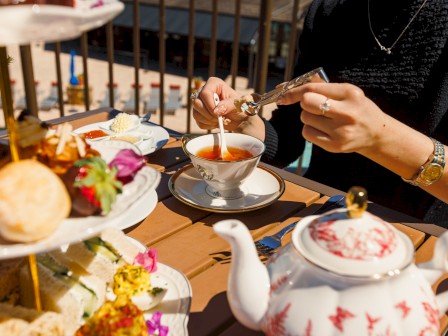 A person at a tea party is dipping treats into tea; there’s a cupcake stand, a teapot, and a cup with a saucer on a wooden table.