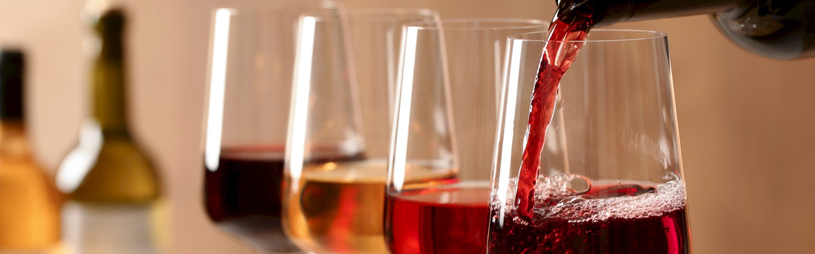 A bottle pouring red wine into one of four glasses, with more wine bottles in the background.