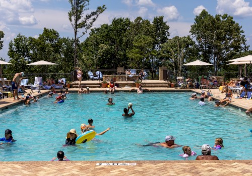 People enjoying a sunny day at a crowded outdoor swimming pool with trees and umbrellas in the background.