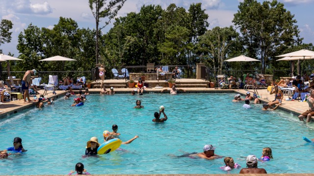 People enjoying a sunny day at a crowded outdoor swimming pool with trees and umbrellas in the background.