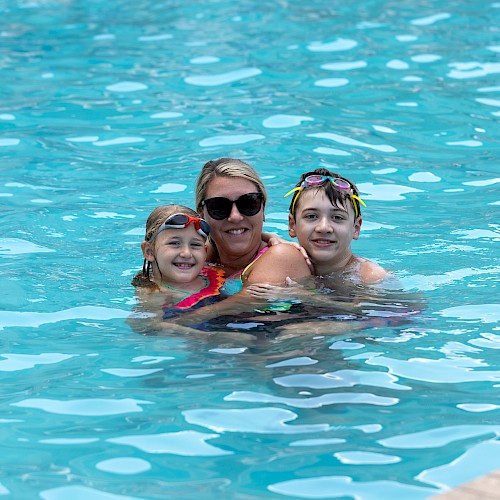 A woman with two children enjoy swimming and smiling in a bright blue pool on a sunny day.