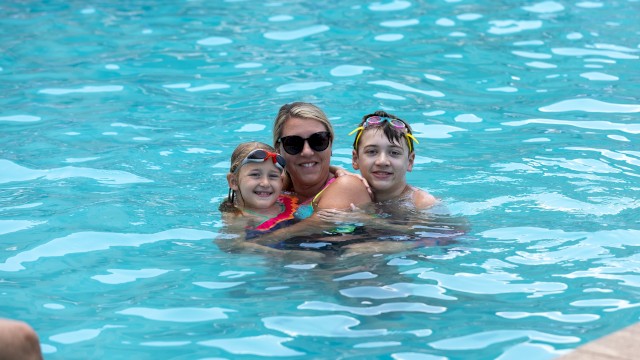 A woman with two children enjoy swimming and smiling in a bright blue pool on a sunny day.
