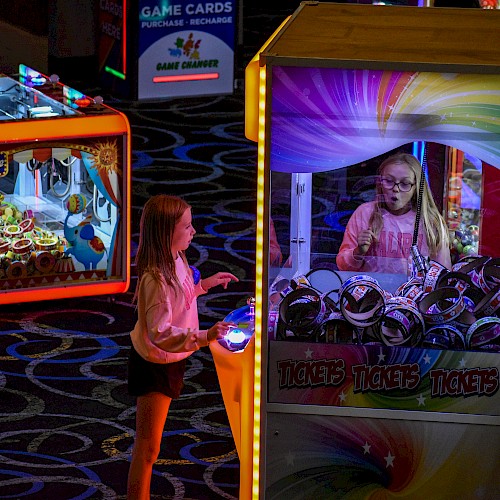 An arcade with two girls playing claw machines filled with toys, vibrant lights, and patterned carpet.