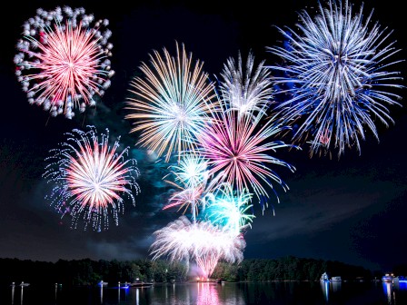A display of colorful fireworks bursting in the night sky above a lake, with reflections in the water and a silhouetted treeline.